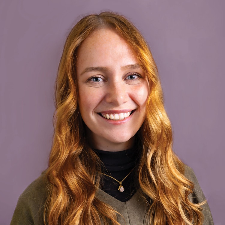 A young woman with long, wavy red hair smiles broadly, wearing a black turtleneck and green top.