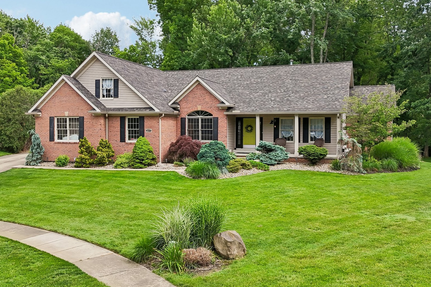 A brick house with a gray roof, black shutters, and a bright yellow door, surrounded by green trees and a lawn.