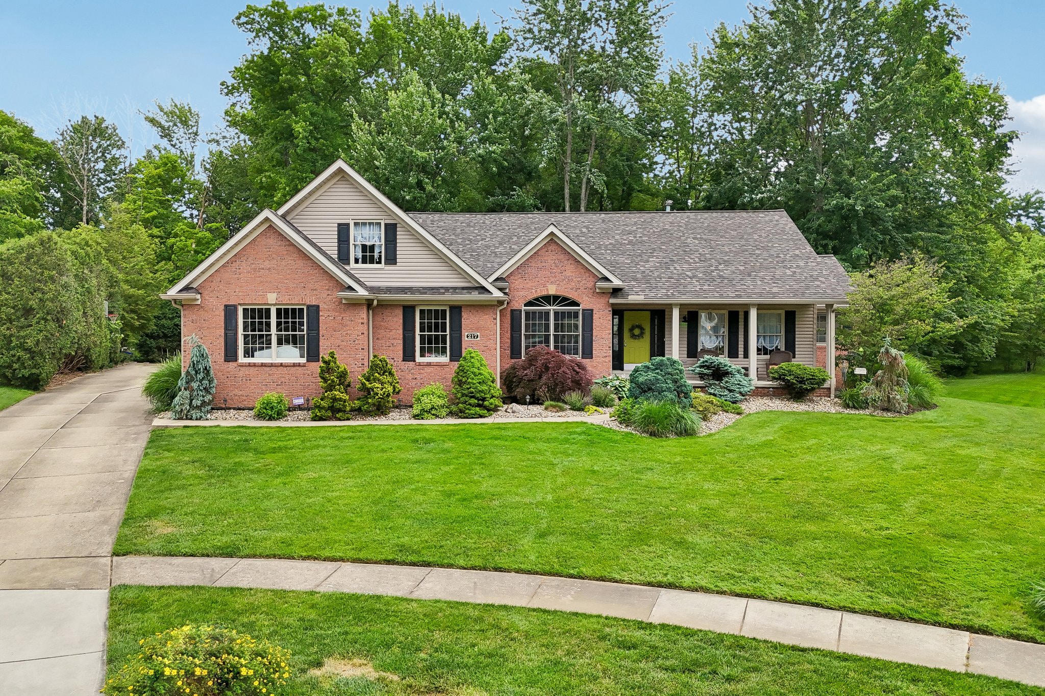 A brick house with a gray roof and a bright yellow door, surrounded by green trees and a lawn.