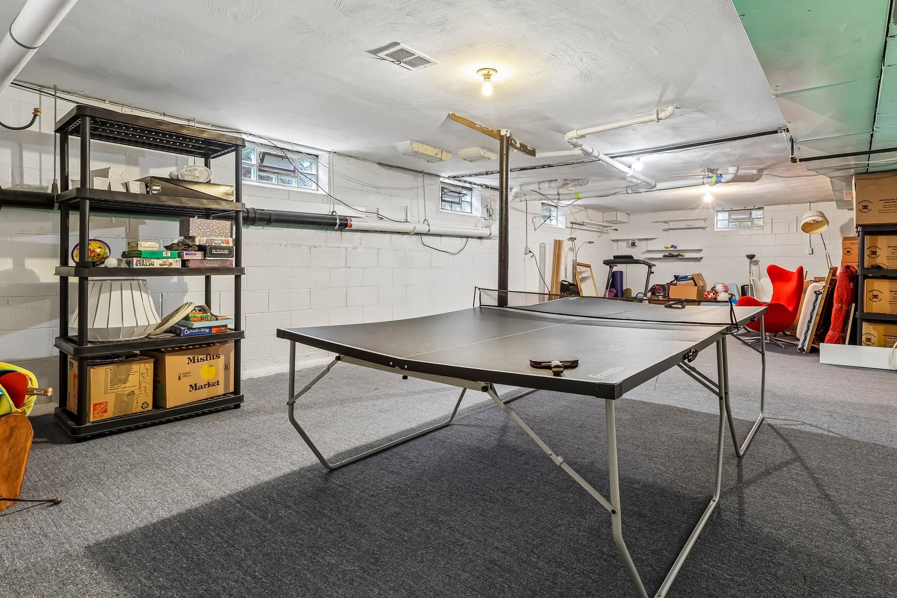 A basement with a black ping-pong table, a black shelf with boxes and games, and a red chair.